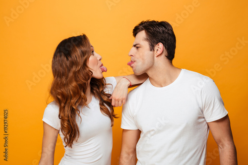 Portrait of funny man and woman in white t-shirts smiling and looking on each other with tongues sticking out, isolated over yellow background