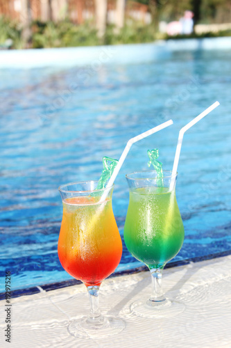 Two glasses with colored cocktails are around the pool