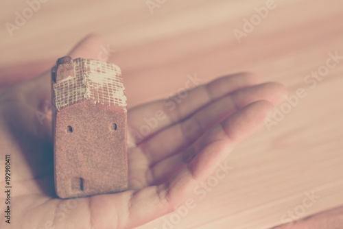 hand holding brown ceramic house model, blurred wooden pallet as background, selective focus, dream of home concept