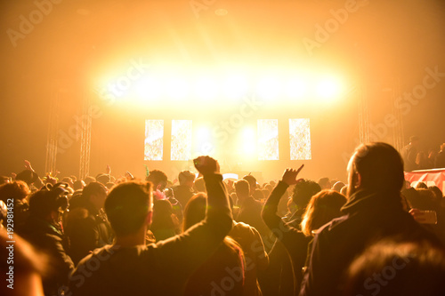 People with hands up during a concert in a hangar. Led wall and orange lights. Laser and lights illuminate the party. View of the stage from the dance floor.Dj on the stage