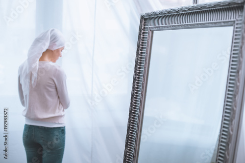 back view of young sick woman in kerchief standing near mirror, cancer concept