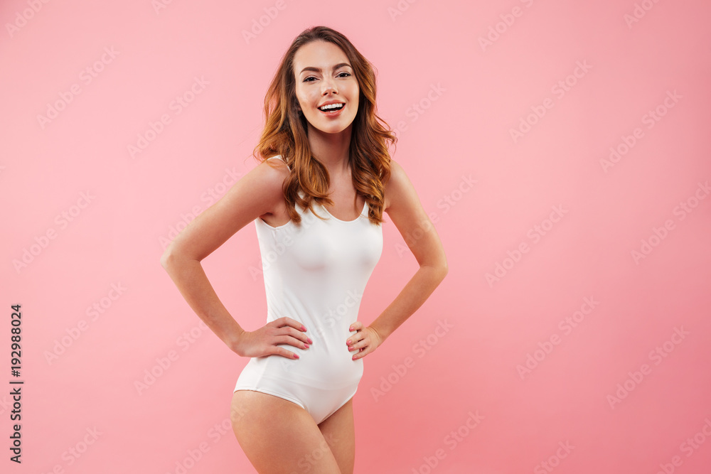 Caucasian sexual woman with brown hair wearing one-piece swimsuit smiling and posing on camera keeping hands on waist isolated over pink background Caucasian sexual woman with brown hair wearing one-piece swimsuit smiling and posing on camera keeping hands on waist isolated over pink background
