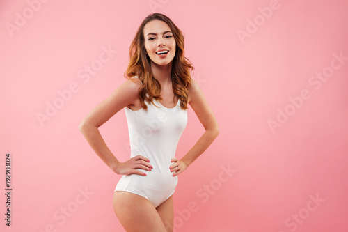 Caucasian sexual woman with brown hair wearing one-piece swimsuit smiling and posing on camera keeping hands on waist isolated over pink background