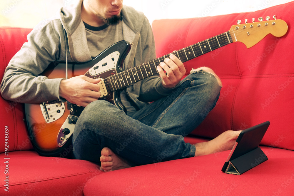 Obraz premium Casually dressed young man with guitar playing songs in the room at home. Online guitar lessons concept. Male guitarist practicing chord grips.