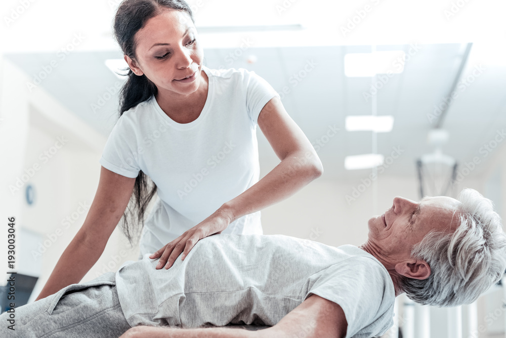 Fototapeta premium Training together. Vigorous old wrinkled grey-haired man lying on a ball for exercises and a joyful young dark-haired afro-american woman helping him and touching his stomach