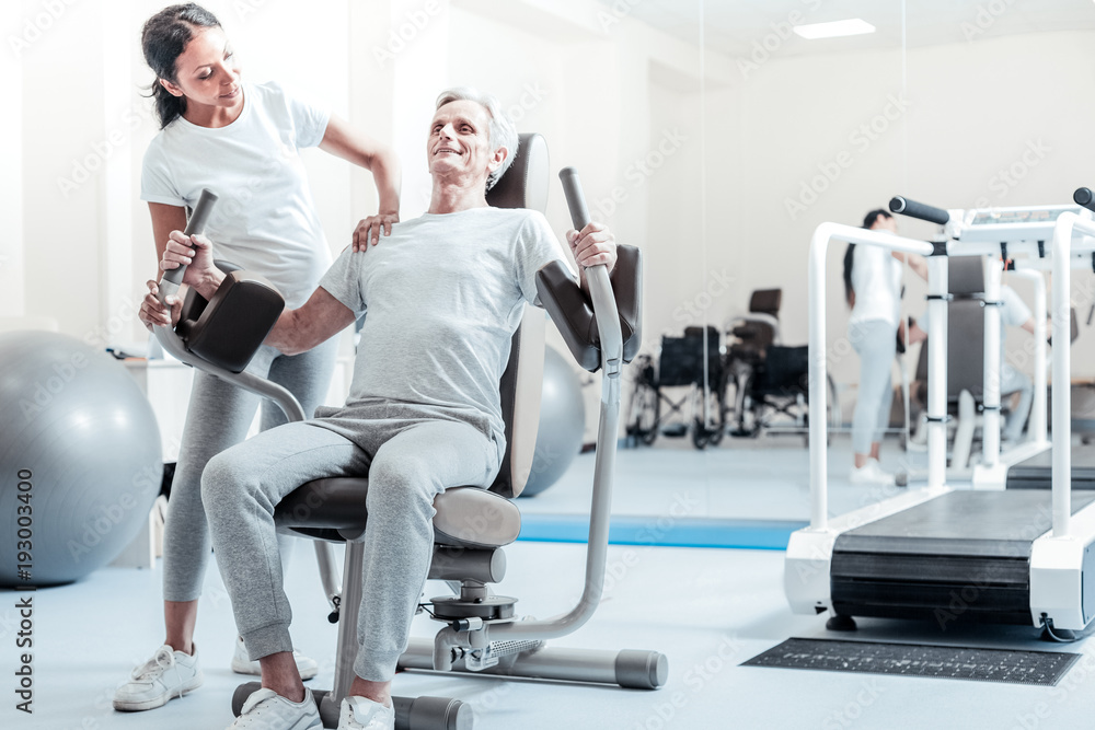 Healthy lifestyle. Happy old grey-haired man smiling and exercising on a training device and a pretty young dark-haired woman standing near him and touching him on the shoulder Healthy lifestyle. Happy old grey-haired man smiling and exercising on a training device and a pretty young dark-haired woman standing near him and touching him on the shoulder