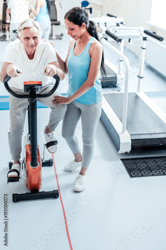 Fototapeta Naklejka Na Ścianę i Meble -  Doing sport. Happy smiling aged grey-haired man exercising on a training device and training his legs while a beautiful smiling young dark-haired female trainer helping and standing near him