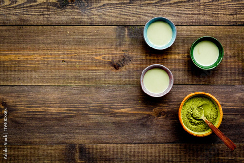 Brew matcha tea. Bowl with powder and cups with beverage on dark wooden background top view copy space