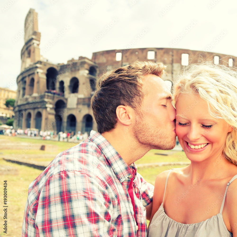 Love - Couple kissing having fun in Rome by the Colosseum. Romantic tourists on holidays vacation travel and man kissing woman on cheek. Beautiful blonde girl and guy in 20s. Coliseum, Rome, Italy.