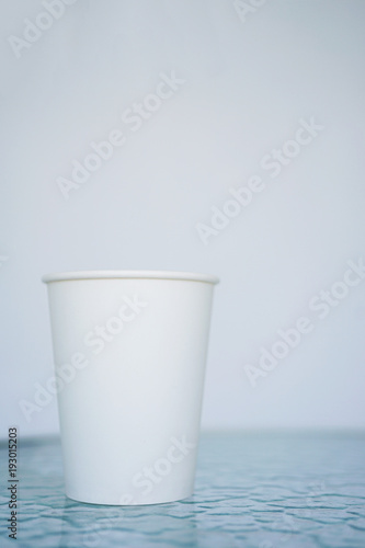 A white paper glass (one-time) for coffee is on the table. Background is blurred, white and blue. Place for the title on the glass. Side view with copy space