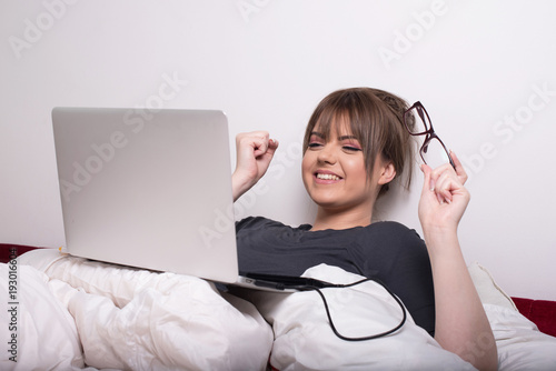 young woman in bed looking at laptop with smile on her face photo