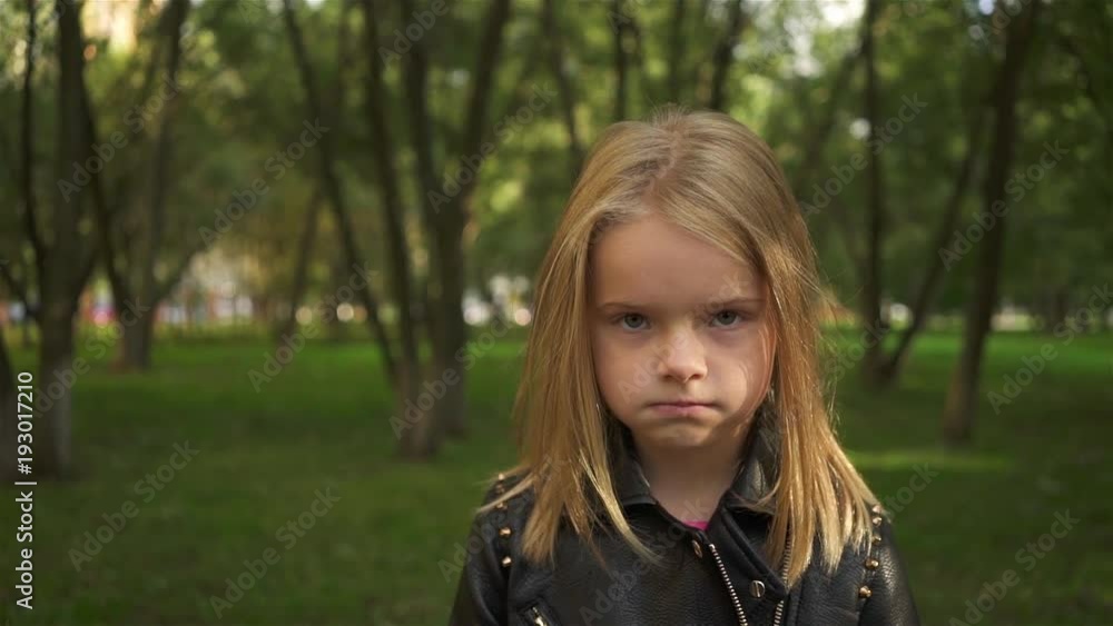 Serioius little blonde girl wearing a leather jacket is fed up. She is standing in a park on a summer day. Handheld slow motion establishing shot Serioius little blonde girl wearing a leather jacket is fed up. She is standing in a park on a summer day. Handheld slow motion establishing shot