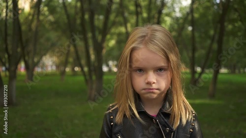 Serioius little blonde girl wearing a leather jacket is fed up. She is standing in a park on a summer day. Handheld slow motion establishing shot photo