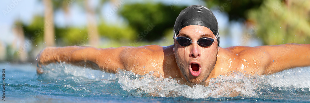 Man swimmer swimming butterfly strokes in pool competition training for triathlon. Professional male sport athlete swimmer wearing swimming goggles and cap coming out of water portrait.
