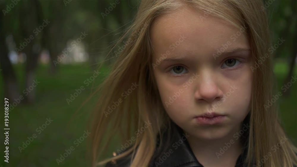 Serious and cute blonde girl wearing a leather jacket looking at camera standing in a park. Handheld slow motion close up shot Serious and cute blonde girl wearing a leather jacket looking at camera standing in a park. Handheld slow motion close up shot