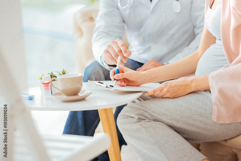 Filling medical form. Scaled up look on an expectant mother sitting next to a doctor and writing something down. Filling medical form. Scaled up look on an expectant mother sitting next to a doctor and writing something down.