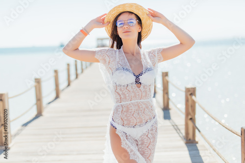 Girl pose at sea pier in straw hat and sunglasses. Woman in sexy swimsuit on tropical beach on sunny blue sky. Summer vacation. Recreation, activity, leisure concept.