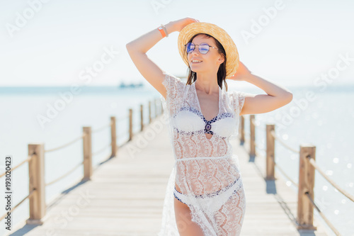 Young beauty smile woman in white bikini, on hat and sunglasses possing on pier on sea background