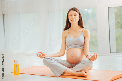 Minute of meditation. Calm deep pregnant woman sitting in the spacious room on the gymnastic rug holding eyes closed and mediating.