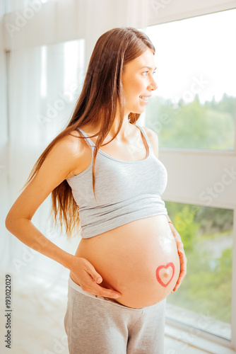 Free Download Real happiness. Joyful pregnant satisfied woman standing in the bright room touching to stomach and looking through the window. - GFXLoad
