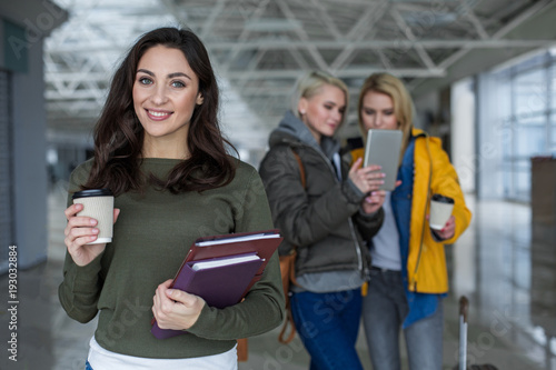 Waist up portrait of beautiful lady with happy face holding cup of drink and literature. Focus on her. Two blonde haired women using tab on background