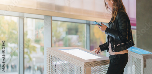 Young woman stands indoor and touches digital display while holding smartphone in her hand. Hipster girl is connected to cloud technology. Innovative technologies, digital display, urban navigation.