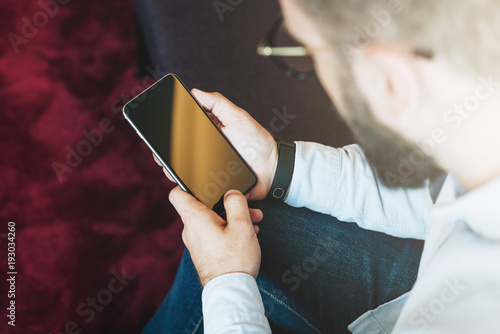 Back view. Young bearded businessman in glasses sits and uses smartphone. Close-up of smartphone with blank screen in male hands. Guy works online. Man blogging, chatting, browsing internet, reading.