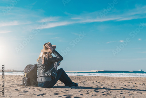 Back view. Young woman tourist in hat and with backpack sitting on beach, using smartphone, looking at sea, on coastline, on horizon. Tourism, vacation, lifestyle. Hipster girl resting on beach. © foxyburrow