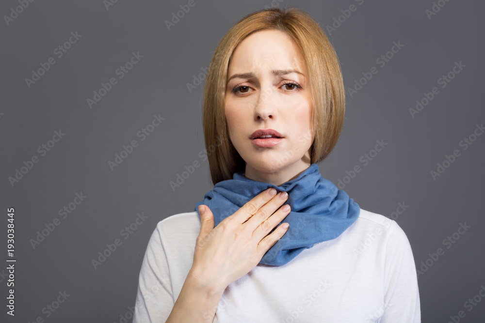 Portrait of ill blonde female touching her throat and looking at camera with sadness, her neck wrapped with scarf. Copy space in left side. Isolated on background Portrait of ill blonde female touching her throat and looking at camera with sadness, her neck wrapped with scarf. Copy space in left side. Isolated on background
