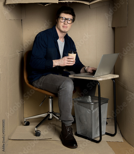 Feeling uncomfortable concept. Full length portrait of manager is sitting at table in cramped carton office while drinking coffee and working on modern laptop. He is looking at camera disappointedly photo