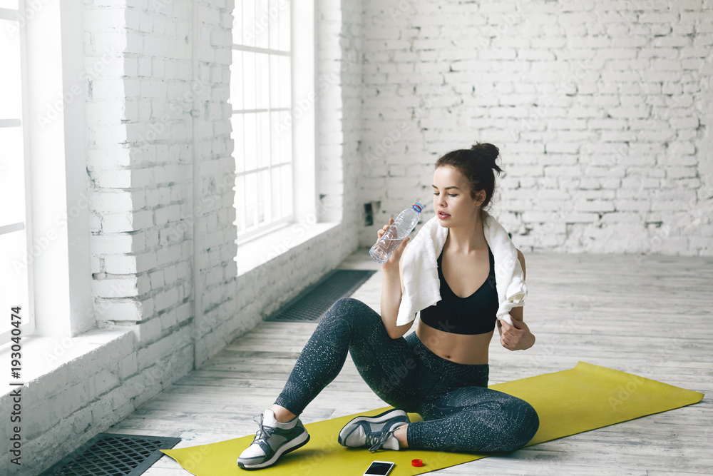 Athletic cute girl in black sports bra, leggings and running shoes relaxing  after cardio training routine in gym hall, resfreshing herself with  drinking water and checking email on her smart phone Stock