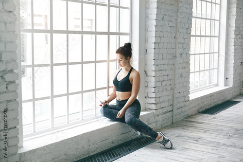 Picture of attractive young woman fitness instructor sitting on windowsill in spacious room before group workout, searching for music tracks on her mobile phone. People, sports and technology