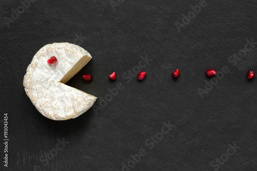 Camembert or brie cheese cut out like pacman eating pomegranate seeds on black slate background