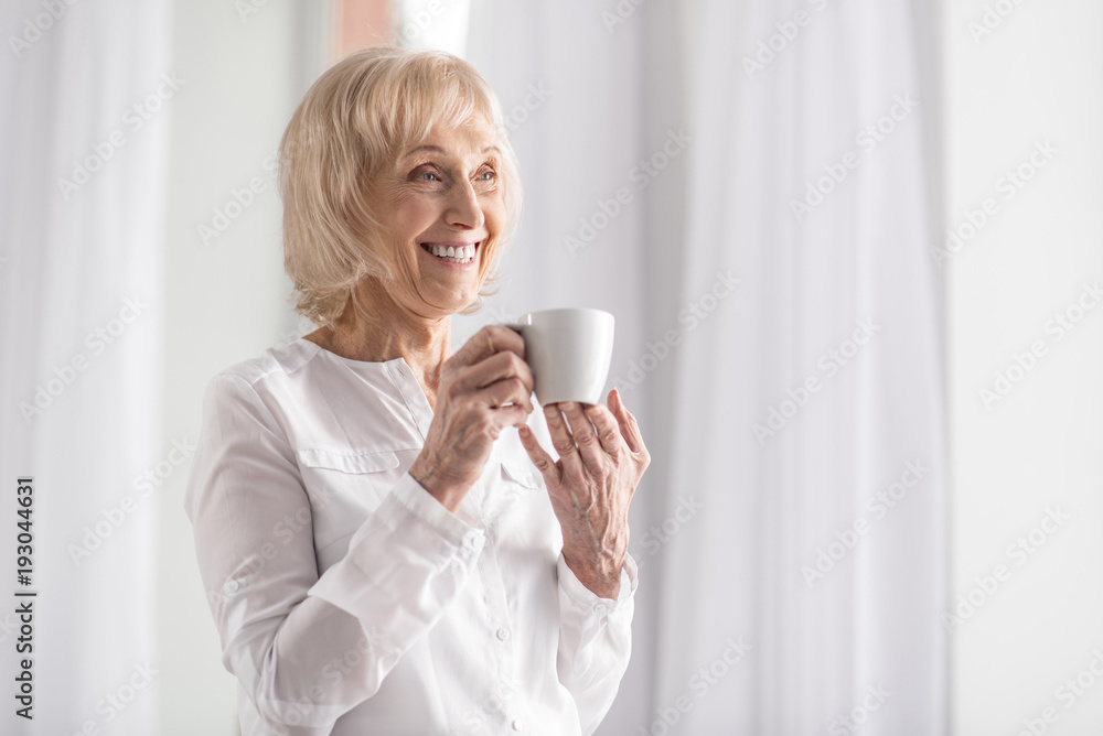 Back coffee. Happy vigorous mature woman laughing while standing on the light background and drinking coffee Back coffee. Happy vigorous mature woman laughing while standing on the light background and drinking coffee