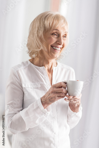 Coffee odor. Gay optimistic mature woman smiling while enjoying coffee and posing on light background