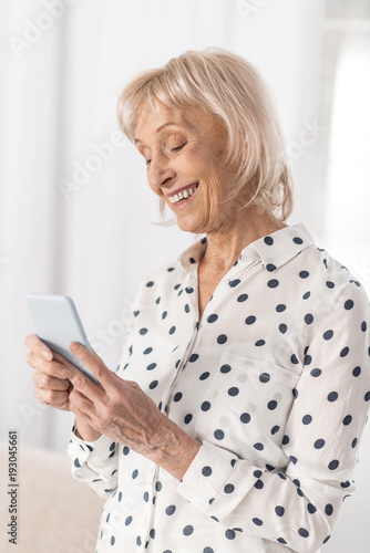Waiting response. Glad elegant mature woman responding message while looking at screen and posing on the light background
