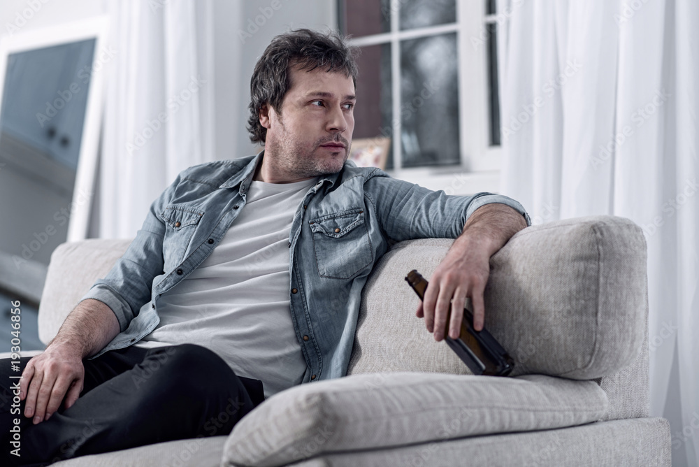 Thoughtful man. Calm unshaved unemployed man sitting on a sofa in his comfortable room and thoughtfully looking to the left while analyzing his condition Thoughtful man. Calm unshaved unemployed man sitting on a sofa in his comfortable room and thoughtfully looking to the left while analyzing his condition