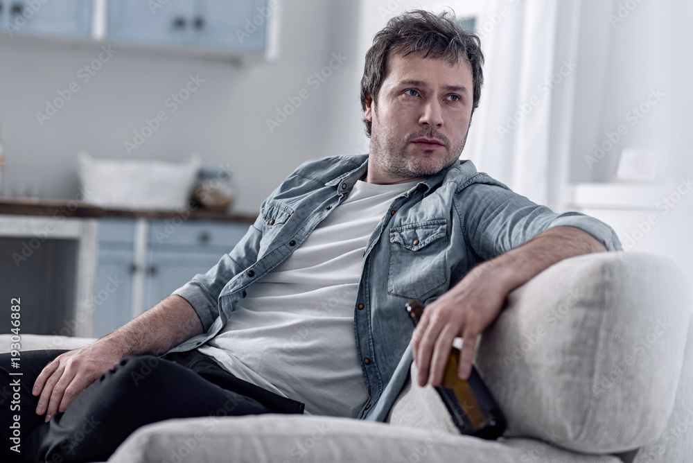 Jobless man. Unshaved lazy man feeling indifferent to life after being unemployed for a long time and sitting on a sofa with a bottle of alcohol in his hand