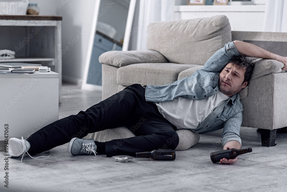 Terrible condition. Unshaved young man resting on the floor with an empty bottle in his hand and feeling unwell after drinking too much alcohol Terrible condition. Unshaved young man resting on the floor with an empty bottle in his hand and feeling unwell after drinking too much alcohol
