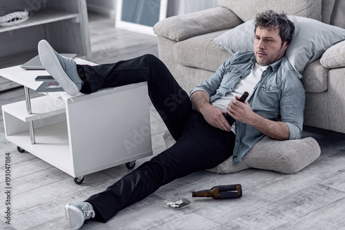 Relaxing alone. Lazy jobless unshaved man looking untidy while resting on the floor with his leg on a little table and drinking alcohol while watching TV