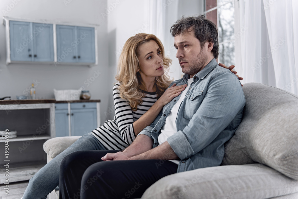 Care. Unhappy young man feeling terrible after losing his job and looking depressed while his loving attentive wife showing her care and support Care. Unhappy young man feeling terrible after losing his job and looking depressed while his loving attentive wife showing her care and support