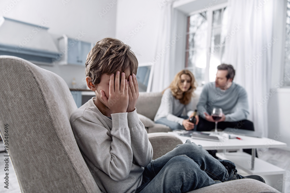 Unhappy childhood. Little emotional scared boy feeling unhappy and closing his face with two hands while his tired serious parents having an unpleasant talk about their problems Unhappy childhood. Little emotional scared boy feeling unhappy and closing his face with two hands while his tired serious parents having an unpleasant talk about their problems