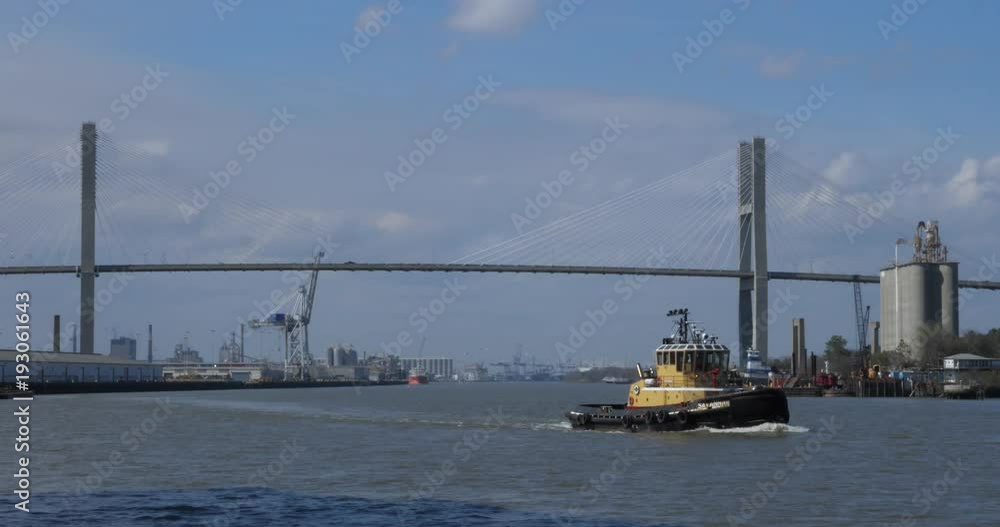 A daytime view of a tug boat traveling on the Savannah River with the Talmadge Memorial Bridge in the distance. A daytime view of a tug boat traveling on the Savannah River with the Talmadge Memorial Bridge in the distance.