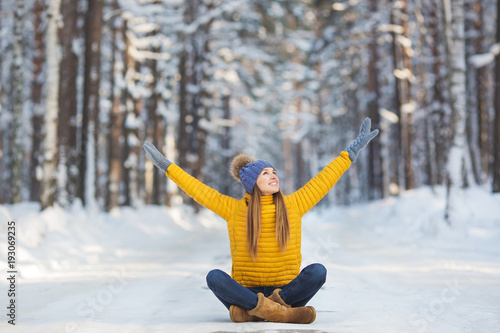 Young smiling woman in a bright clothes sits on a road and hands up in a winter forest