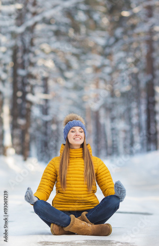 Young smiling woman with closed eyes in a bright clothes sits on a road (yoga) in a winter forest