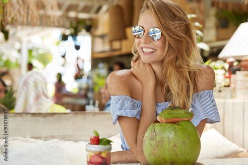 Fashionable young female with bare shoulders, wears stylish summer blouse and trendy shades, admires something with happy expression, drinks coconut cocktail, spends spare time in exotic cafe