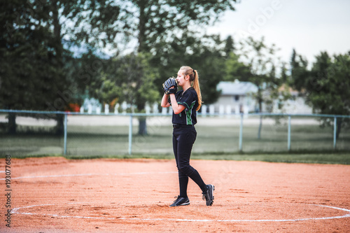 A teenage girl holding glove up to her face with long hair in a ponytail standing on the pitchers mound ready to pitch in a black and green baseball uniform