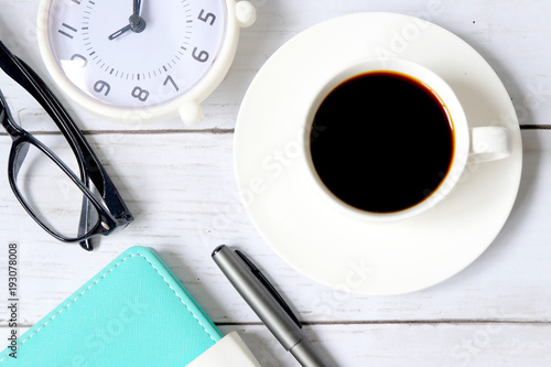 Top view and selective focus of a cup of black coffee with table clock,sunglasses, notebook and pen on white wooden background.