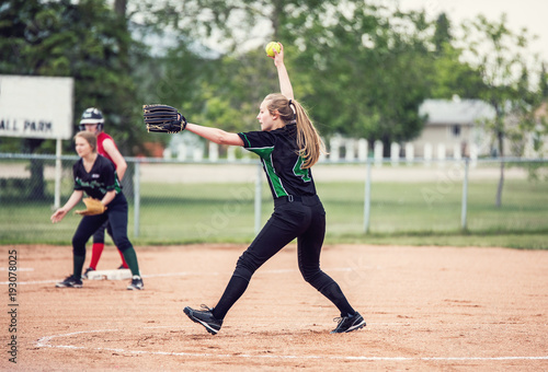A teenaged girl in baseball pitching motion on a pitchers mound with long hair in a ponytail and black and green ball uniform