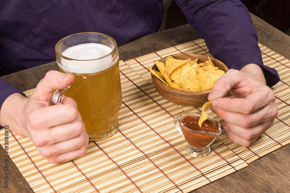Wooden table male hands holding a mug with beer and nachos dipped in the sauce . Copy paste Wooden table male hands holding a mug with beer and nachos dipped in the sauce . Copy paste